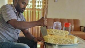 A man sitting on a sofa, cutting a large piece of food on a glass table.