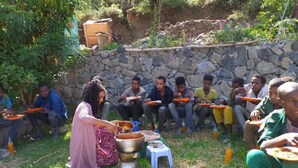 A group of people sitting outdoors in a stone-walled area with a woman serving food.