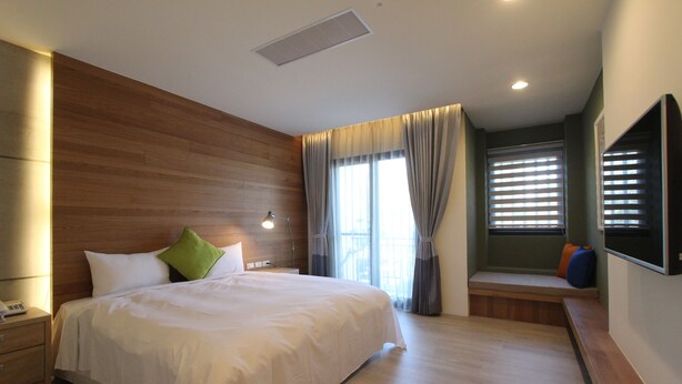 A modern hotel room with a large bed, a bedside table with a lamp, a built-in bench with cushions, and a large window with blinds. - Muguang Inn (Hengchun)