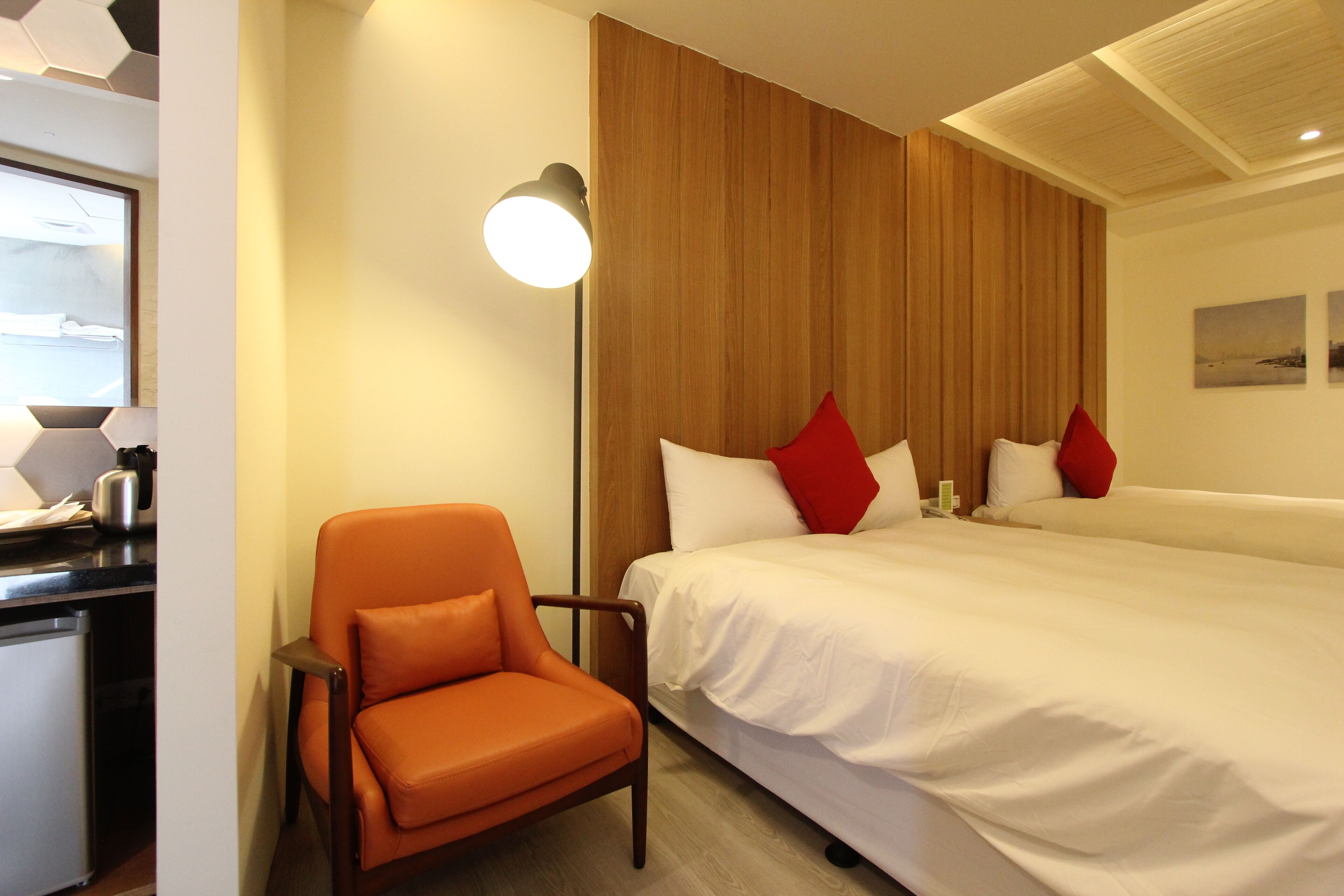 A hotel room with a bed, a chair, a lamp, and a small table with a kettle.