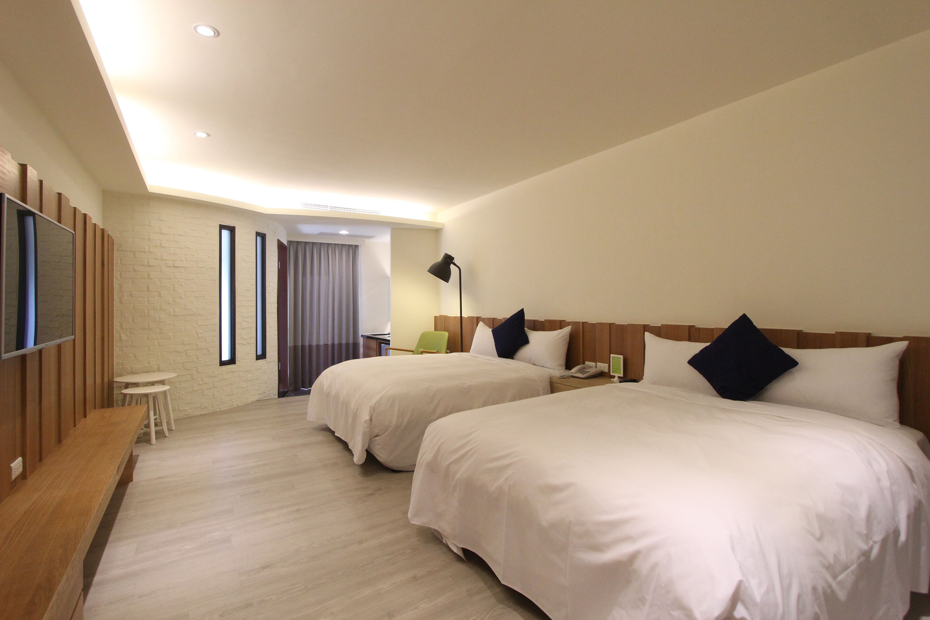 Habitación de hotel con dos camas, un televisor de pantalla plana, un banco de madera, una mesita pequeña y una lámpara.