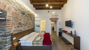 Habitación de hotel con pared de ladrillo, techo de madera, cama con ropa blanca, una manta roja y una mesita de noche con fruta, además de un televisor en la pared.