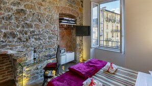 A room with stone walls, a bed with a striped bedspread, a window with a view of buildings, a TV mounted on the wall, and a small table with a bottle and glasses.