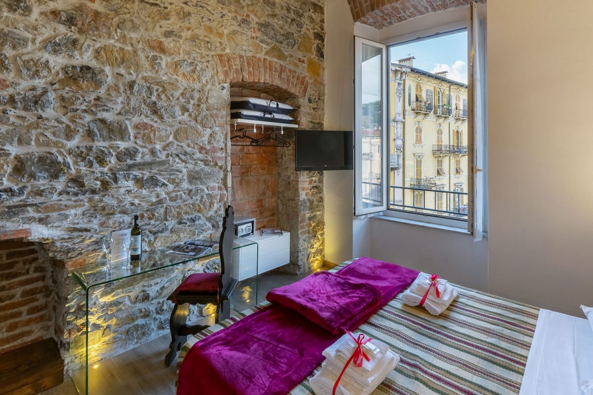 A room with stone walls, a bed with a striped bedspread, a window with a view of buildings, a TV mounted on the wall, and a small table with a bottle and glasses.