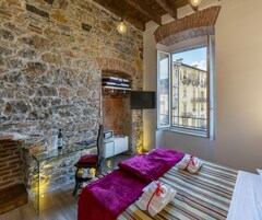 Una habitación con paredes de piedra, una cama con colcha a rayas, una ventana con vistas a edificios, un televisor montado en la pared y una pequeña mesa con una botella y vasos.