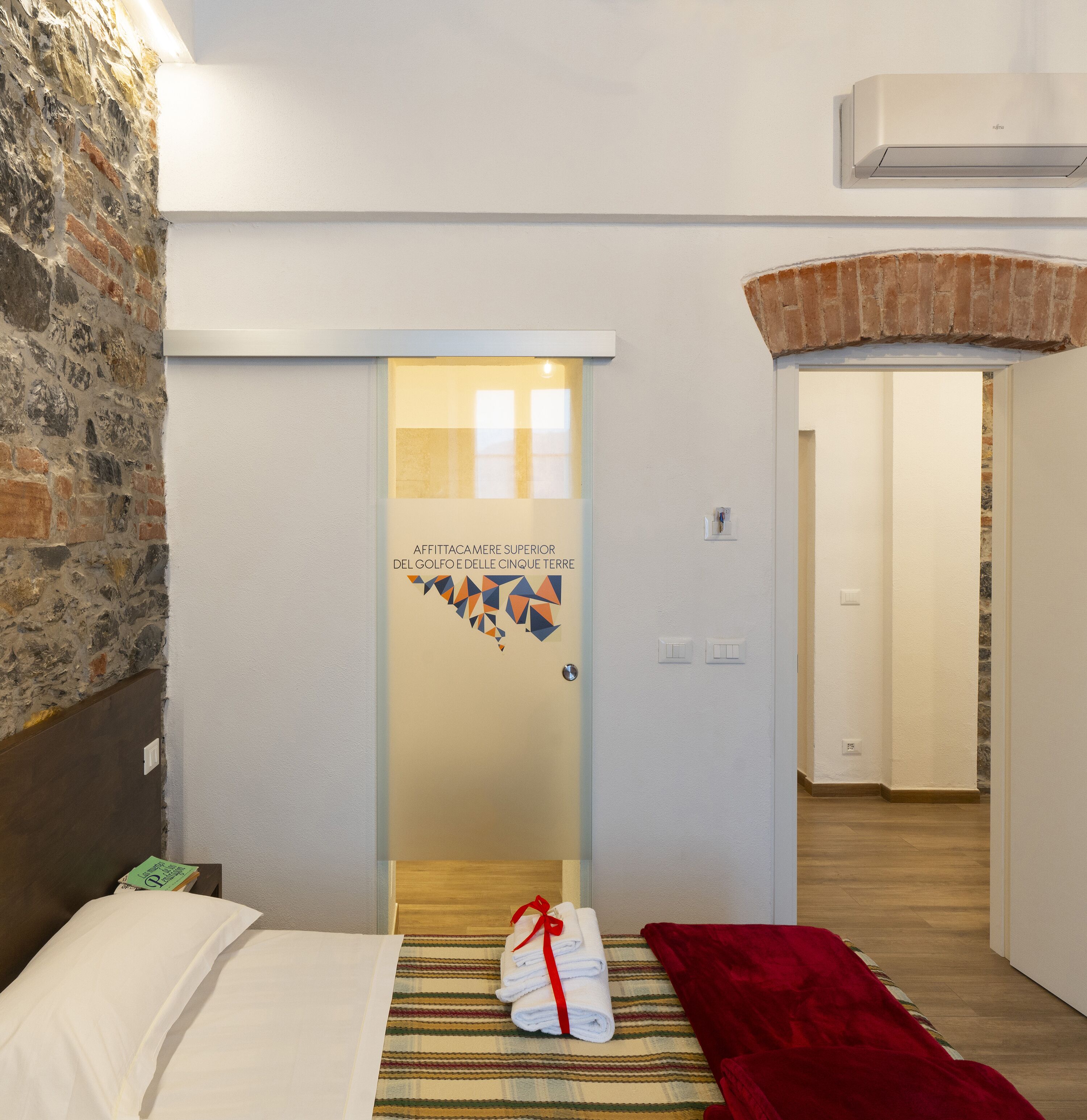 A hotel room with a bed, a red blanket, a striped rug, and a wall with a stone texture.