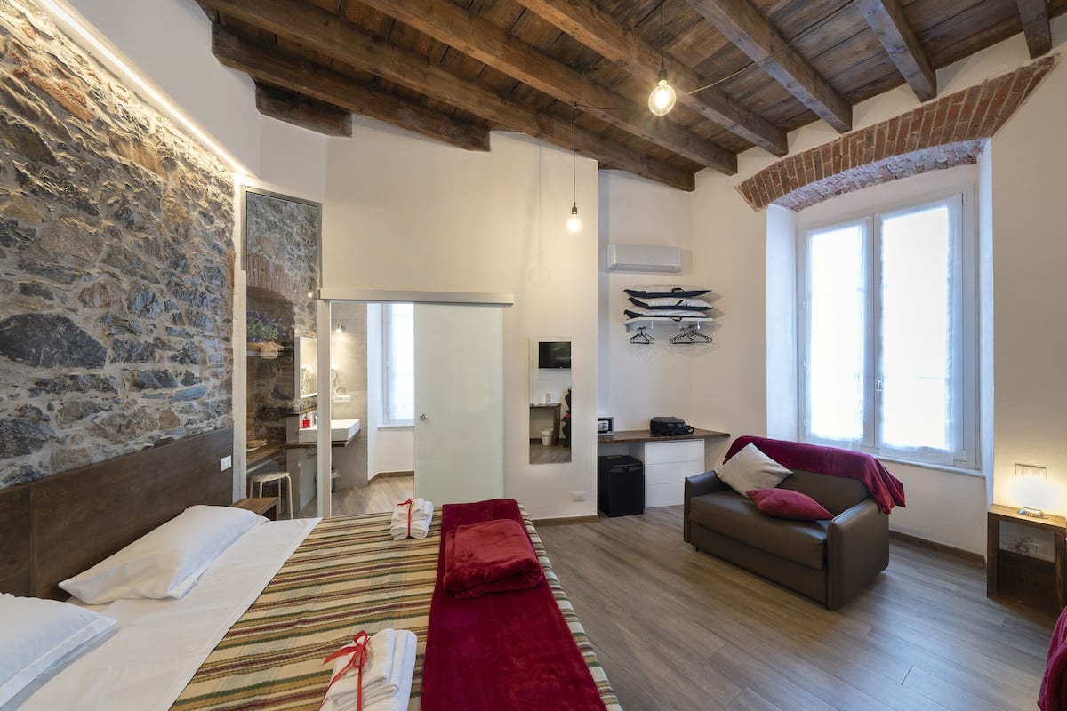 A spacious room with a stone wall, wooden beams, a bed with white linens, a red blanket, a sofa with a red pillow, and a window providing natural light.