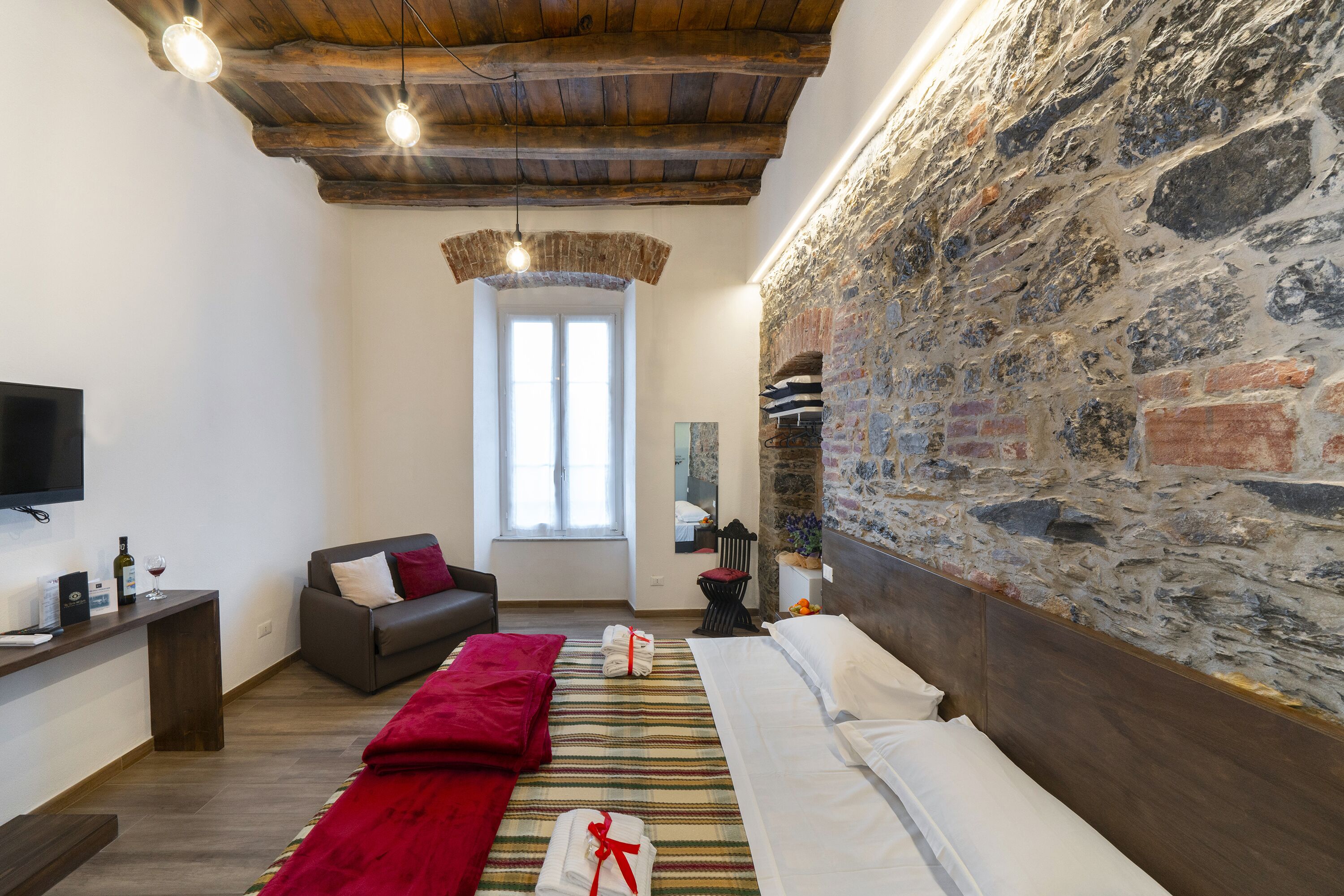 Een knus ingerichte kamer met een stenen muur, houten plafond, een bed met witte lakens, een rode plaid, een grijze bank en een flatscreen televisie op een houten dressoir.