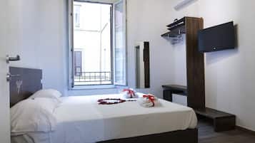 A hotel room with a bed, a TV mounted on the wall, a wooden bench, and a window with a view of the outside.