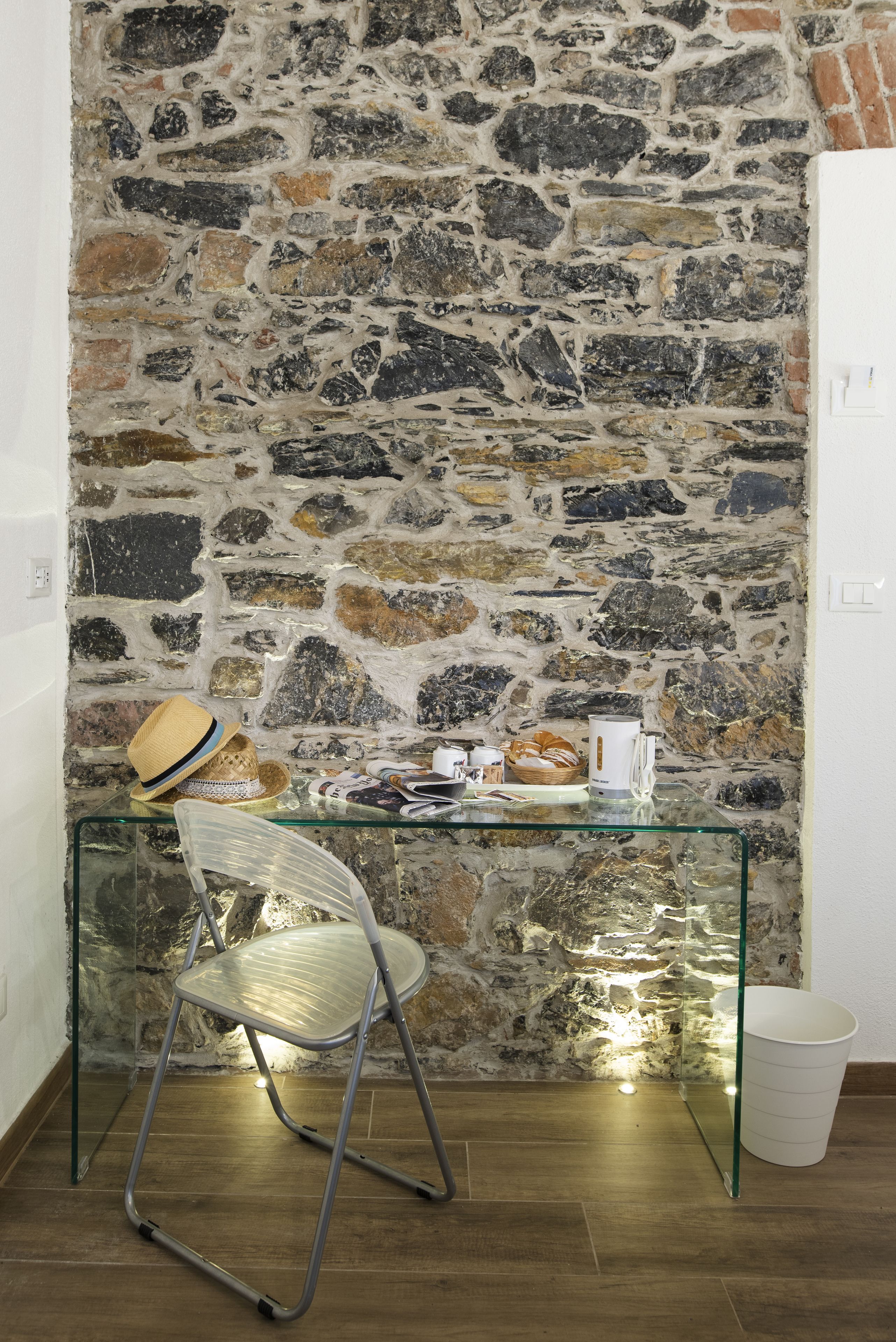 A room with a stone wall, a glass table with various items, a wicker chair, and a white trash bin.