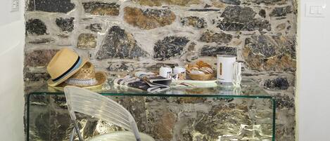 A room with a stone wall, a glass table with various items, a wicker chair, and a white trash bin.