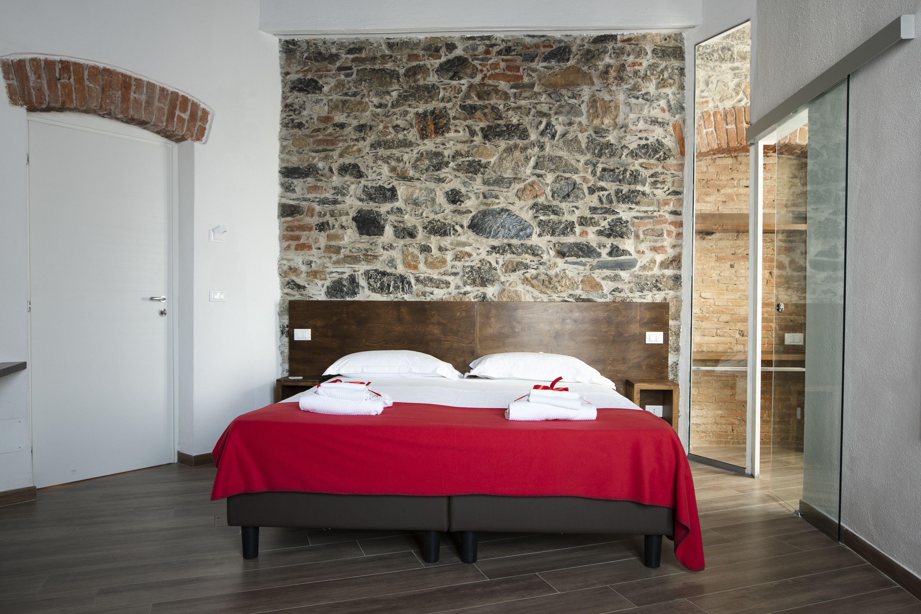 Un lit recouvert d’une courtepointe rouge, avec des oreillers et des serviettes blancs, un mur en pierre en arrière-plan et un plancher en bois.