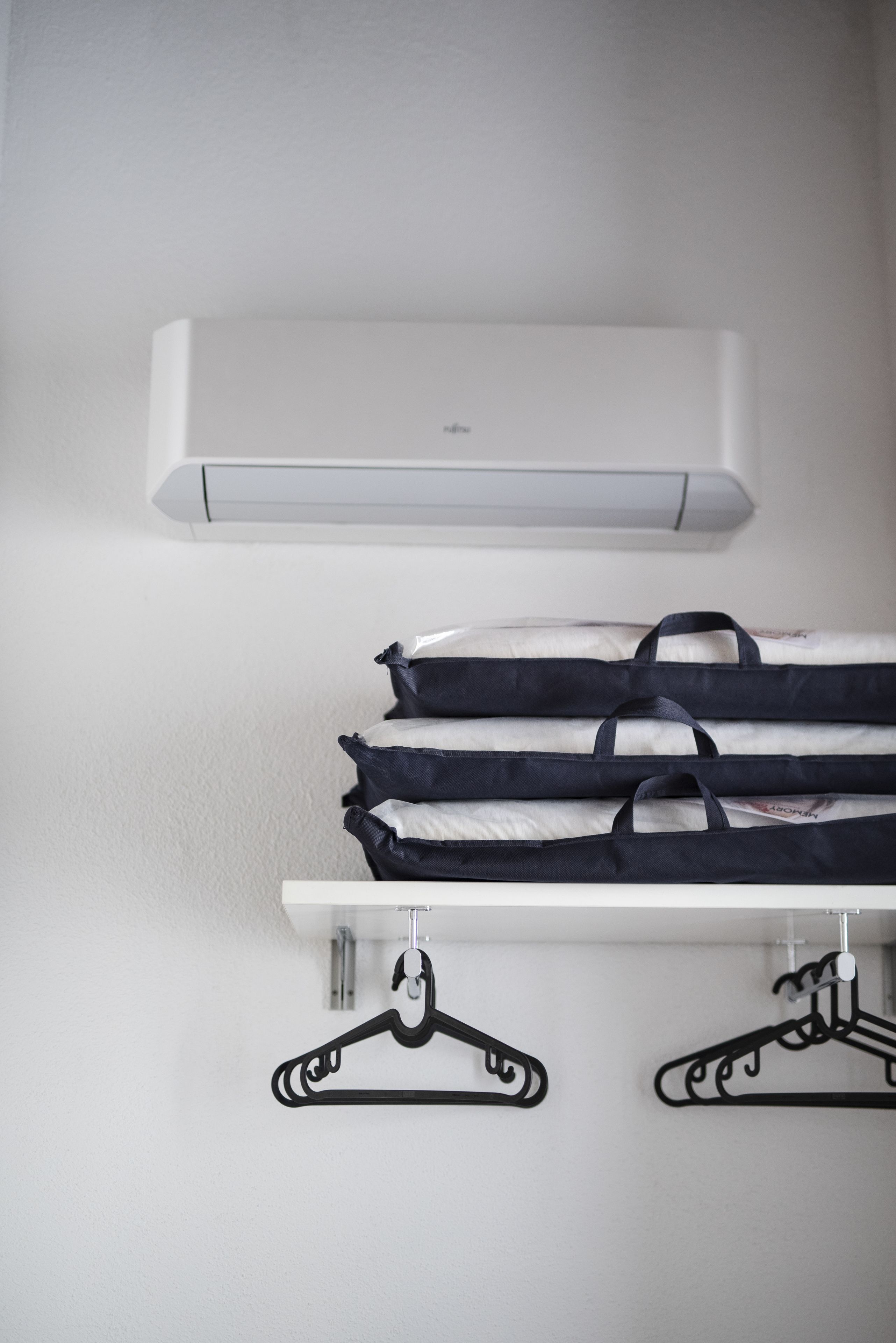 A white wall with a white air conditioner and a white shelf holding folded clothes and two black hangers.