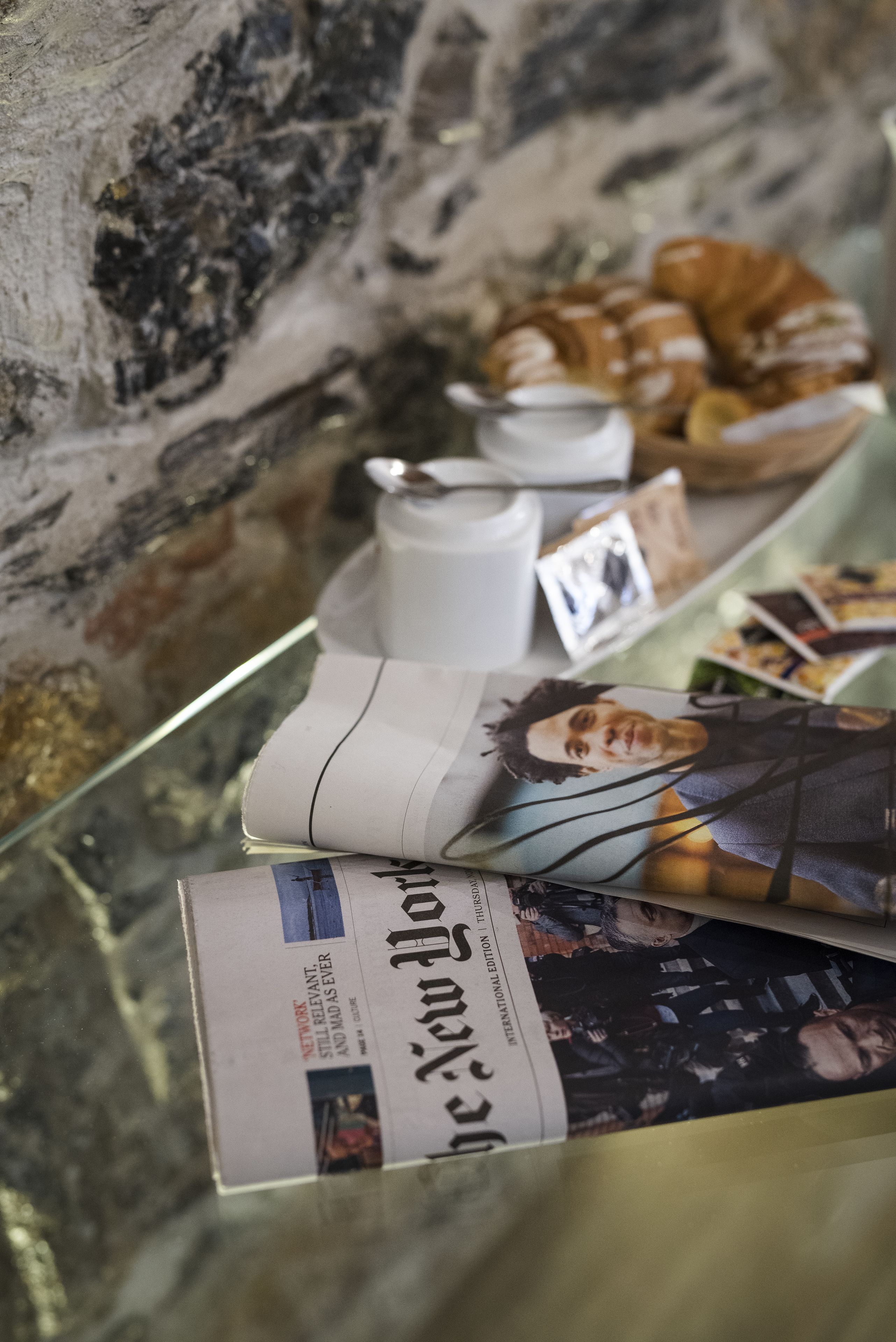A table with a glass top displaying a newspaper, a rolled-up menu, and a few pastries.