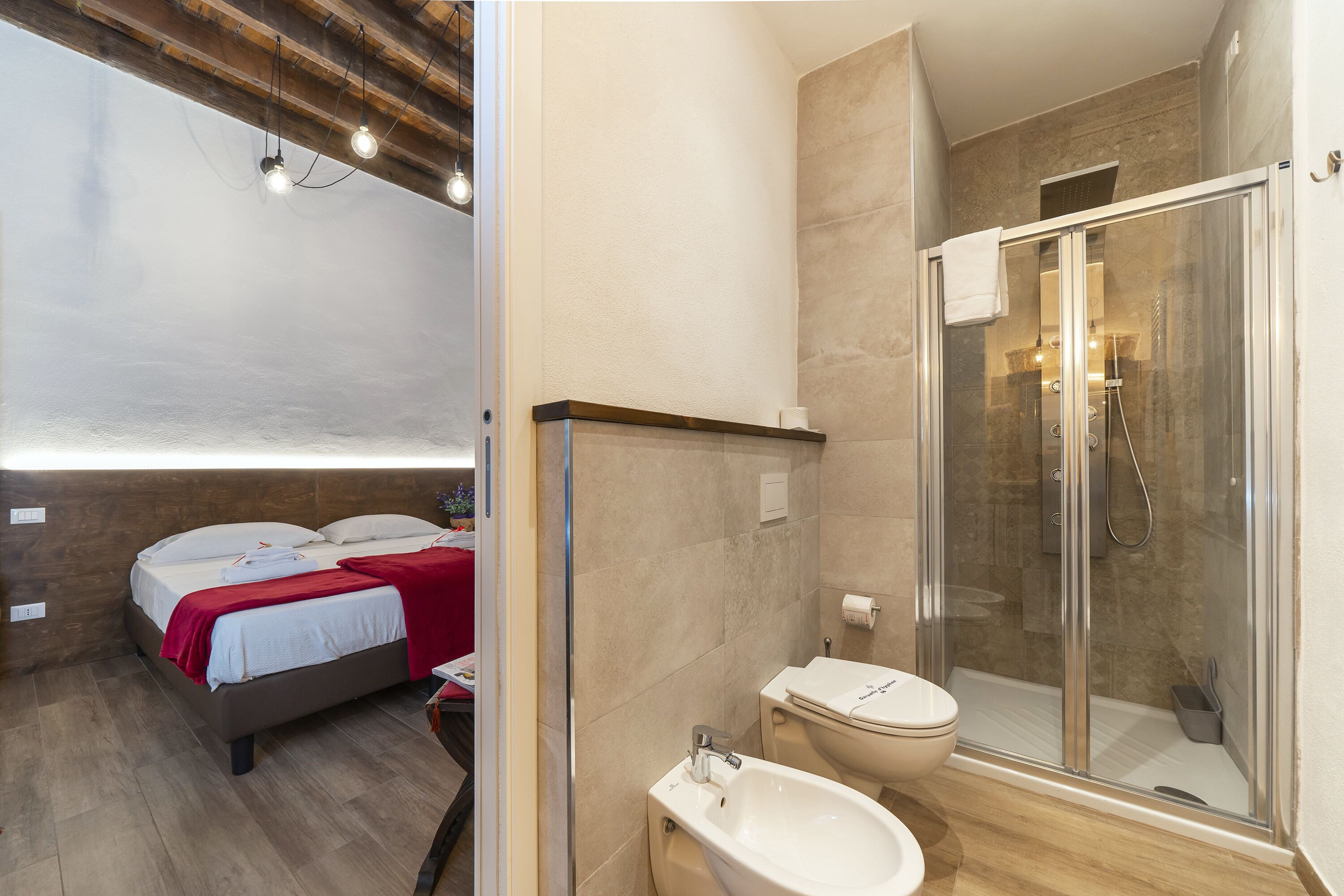 Un dormitorio moderno con cama, mesita de noche y una manta roja. Junto al dormitorio, un baño con cabina de ducha de vidrio, lavamanos e inodoro.