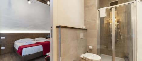 Een moderne slaapkamer met een bed, een nachtkastje en een rode plaid. Grenzend aan de slaapkamer bevindt zich een badkamer met een douchecabine van glas, een wastafel en een toilet.