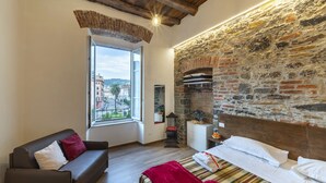 A cozy bedroom with a stone wall, a large window, a bed with a striped bedspread, and a sofa with cushions.