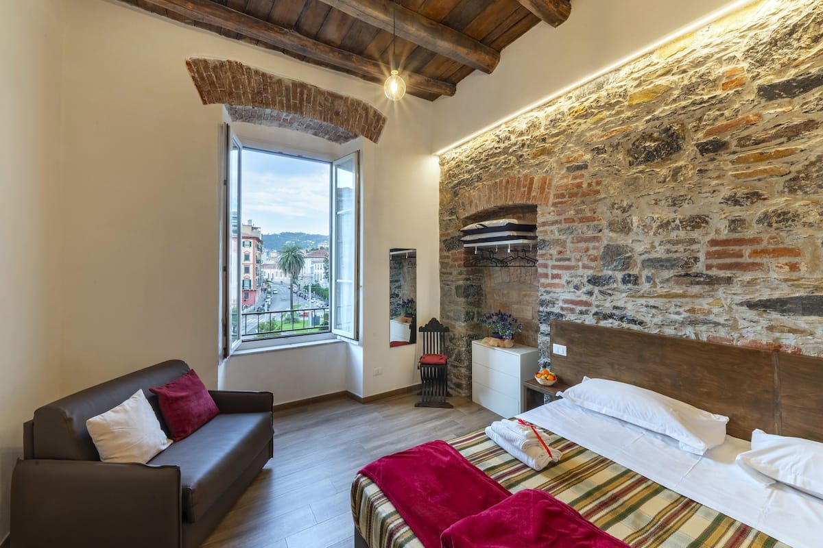 A cozy bedroom with a stone wall, a large window, a bed with a striped bedspread, and a sofa with cushions.
