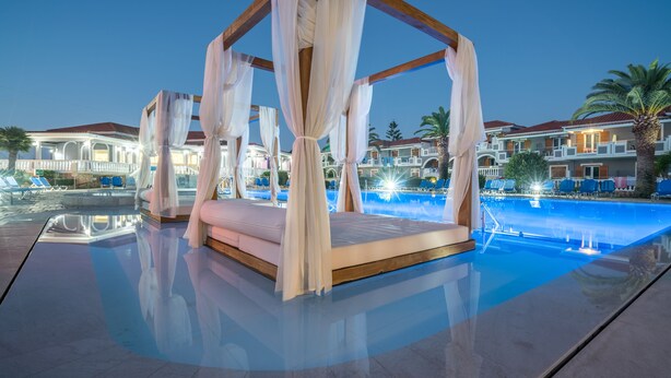 A luxurious outdoor bedroom with a canopy, a pool, and a view of the hotel buildings. - Golden Sun Hotel (Zakynthos)