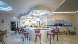A modern bar area with a curved counter, multiple bar stools, pendant lighting, and a view of the city through large windows. - Golden Sun Hotel (Zakynthos)