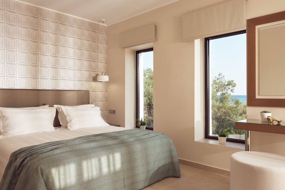 A modern bedroom with a large bed, a view of the sea, and a window with a view of trees.