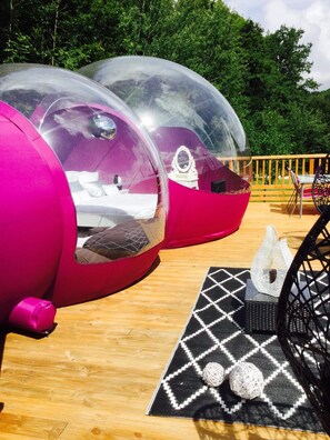 Des modules transparents, en forme de dôme, avec un intérieur rose, disposés sur une terrasse en bois.
