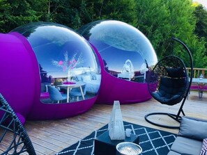 Deux structures sphériques transparentes abritant des sièges à l’intérieur, une chaise suspendue et un cône décoratif sur une terrasse en bois.