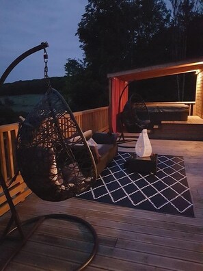 Une terrasse en bois avec un fauteuil suspendu, un canapé et une petite table avec une lampe.