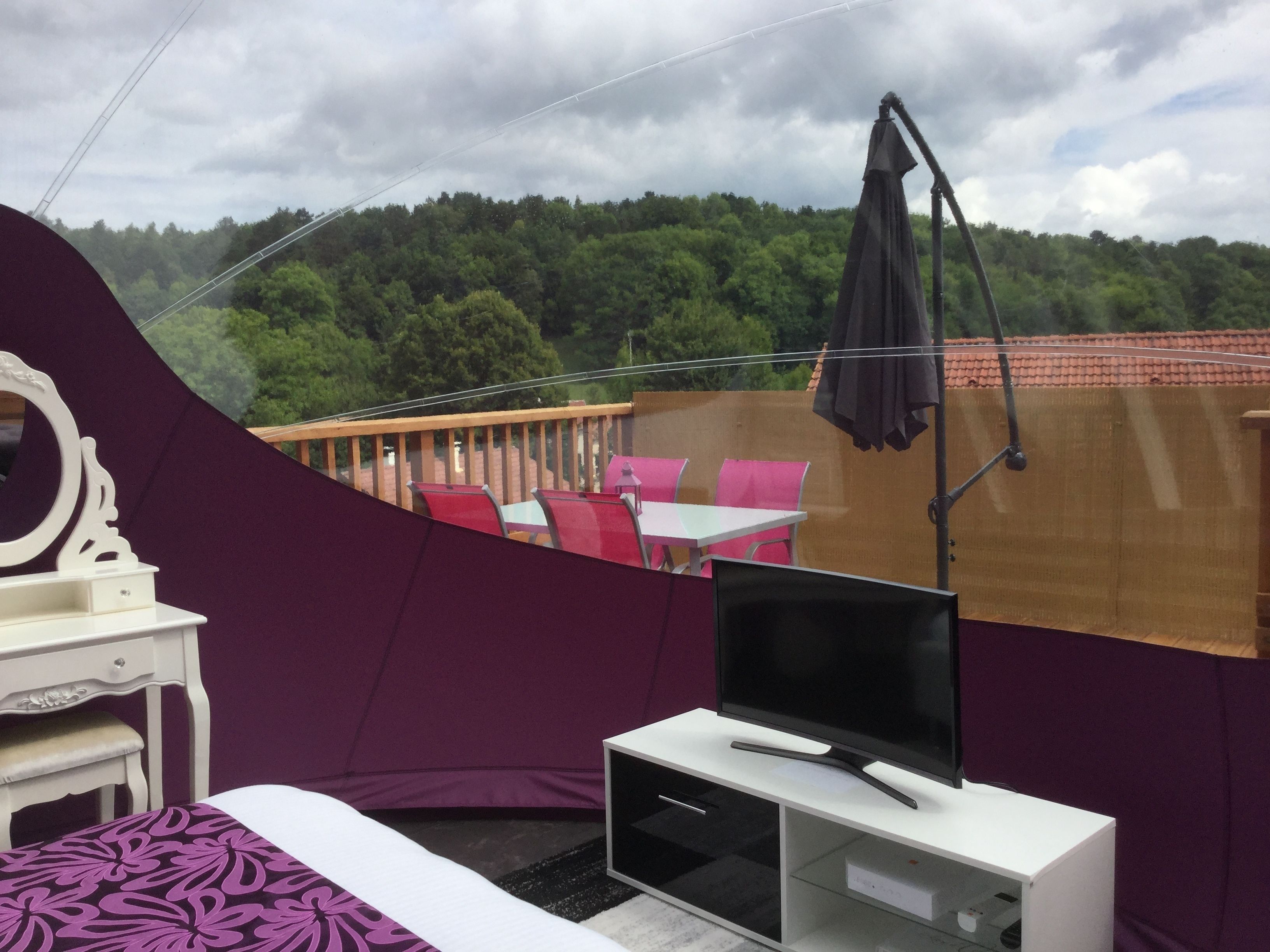 Ein Zimmer mit einem Bett, einem Fernseher auf einer weißen Kommode, einer violetten Wand und Blick auf einen Balkon mit Stühlen und einem Tisch.