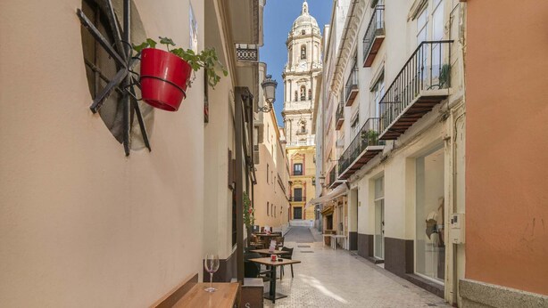 A narrow street with buildings on either side, a red bucket hanging on the left, and a church with a dome in the background.