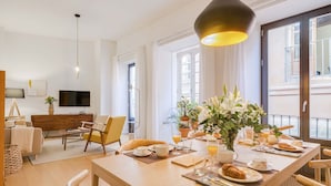 A dining area with a wooden table set for breakfast, a sofa, a TV, and a large window with curtains.