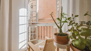 A balcony with a wicker chair, a potted plant, and a view of a building through a window.