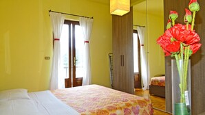 Un dormitorio con cama, espejo y un jarrón con flores rojas.
