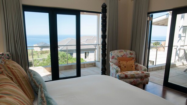 A bedroom with a large bed, a floral armchair, and a view of the sea through floor-to-ceiling windows. - 4 Spinnaker (Knysna)