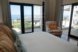 Une chambre avec un grand lit, un fauteuil à motifs floraux et une vue sur la mer grâce à de grandes fenêtres du sol au plafond.