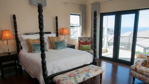 A four-poster bed with a floral bench, two nightstands, a chair, and a view of the sea through large windows. - 4 Spinnaker (Knysna)