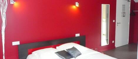 Una habitación con una pared roja, una cama blanca con dos almohadas y una puerta con un espejo.
