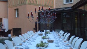 Een lange eettafel gedekt voor een maaltijd, met witte stoelen, een kandelaar en een buffet met broodbakken.