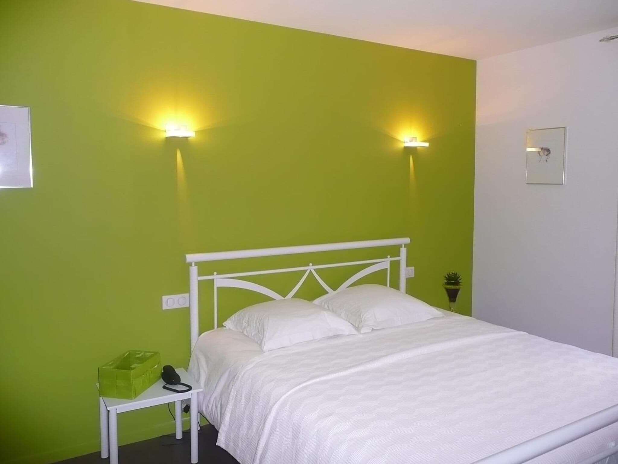 Une chambre avec un lit simple, un cadre en métal blanc, des murs verts et deux lampes fixées au mur.