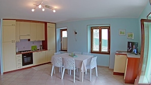 A compact kitchen with a dining area, featuring a table with four chairs, a small refrigerator, and a window with blinds. - Holideal Casa Lauretta (Tremosine)