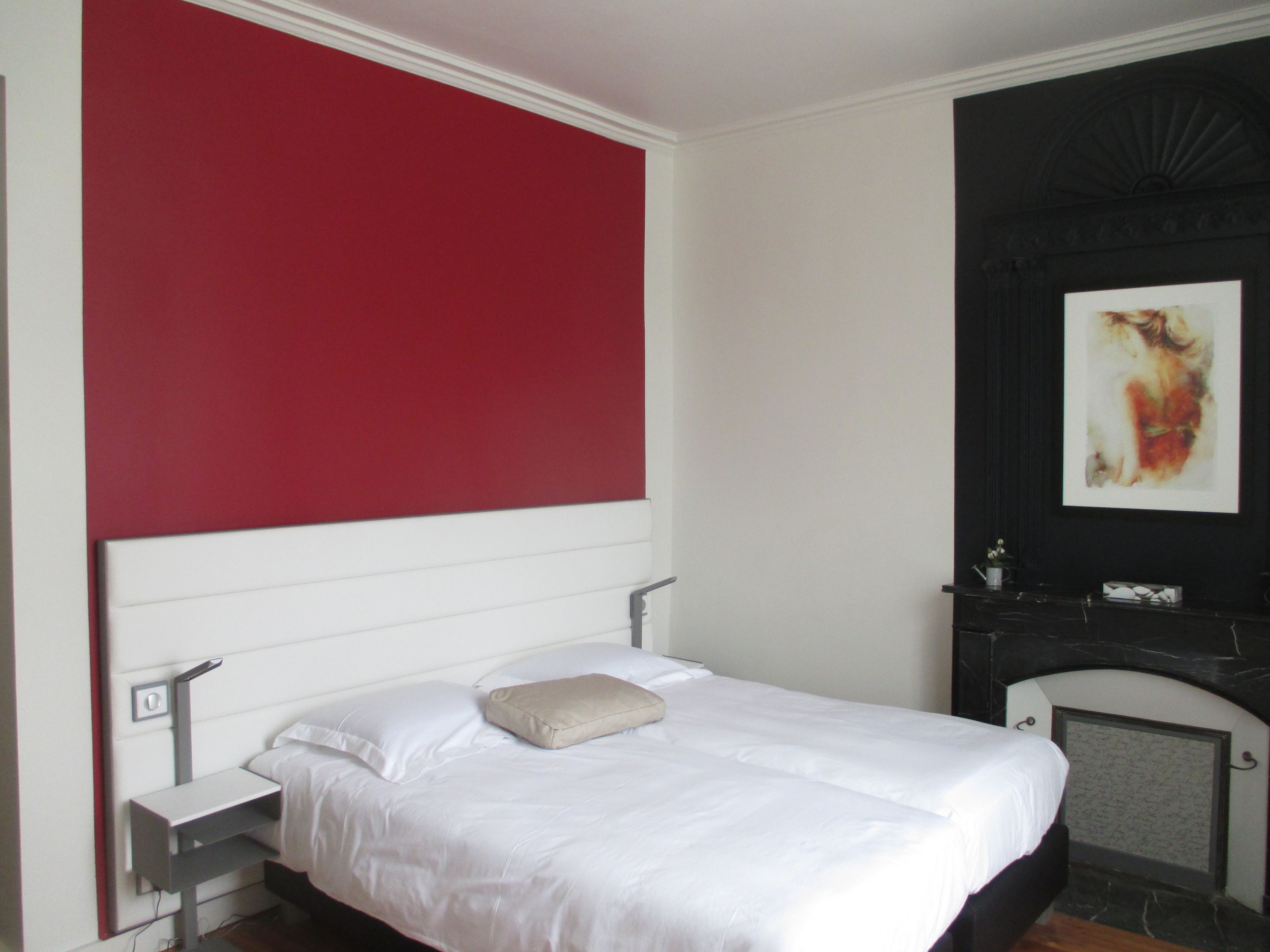 Ein Hotelzimmer mit Bett, Nachttisch, roter Akzentwand und Kamin mit einem Gemälde.