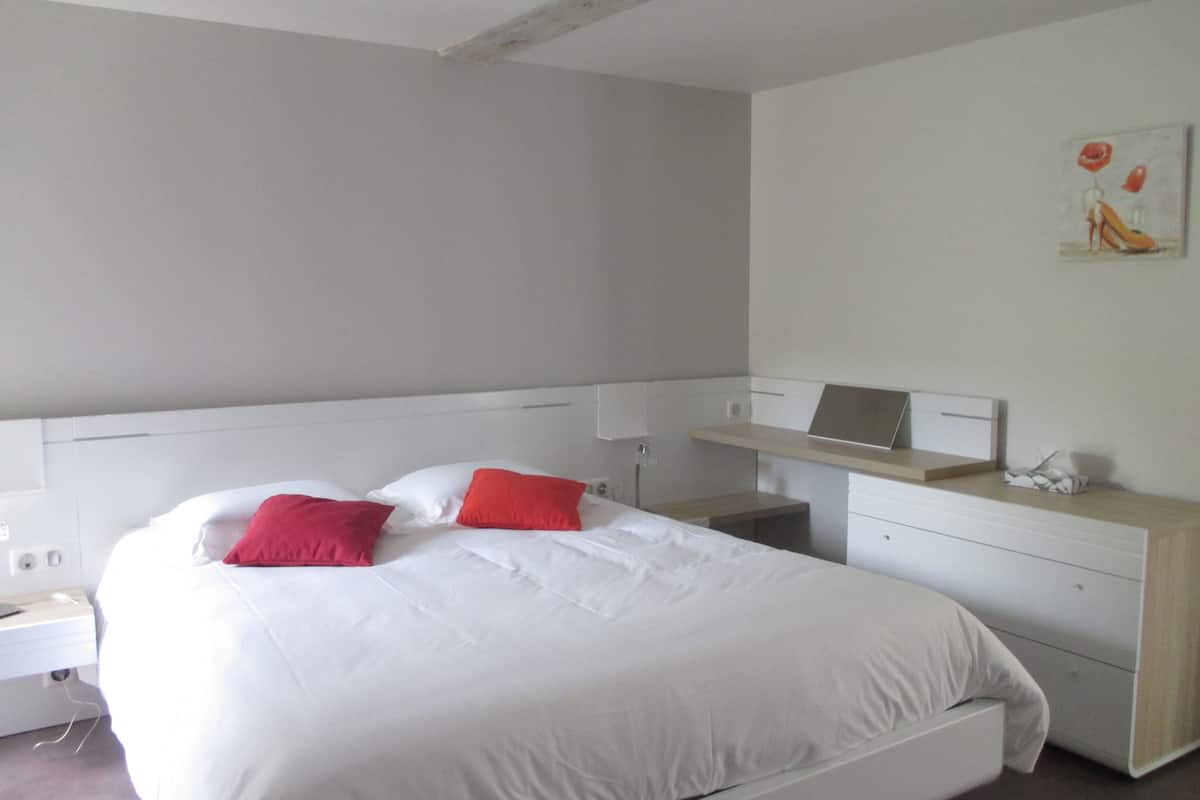 A modern bedroom with a bed, white bedding, a nightstand, a desk with a laptop, and a painting on the wall.