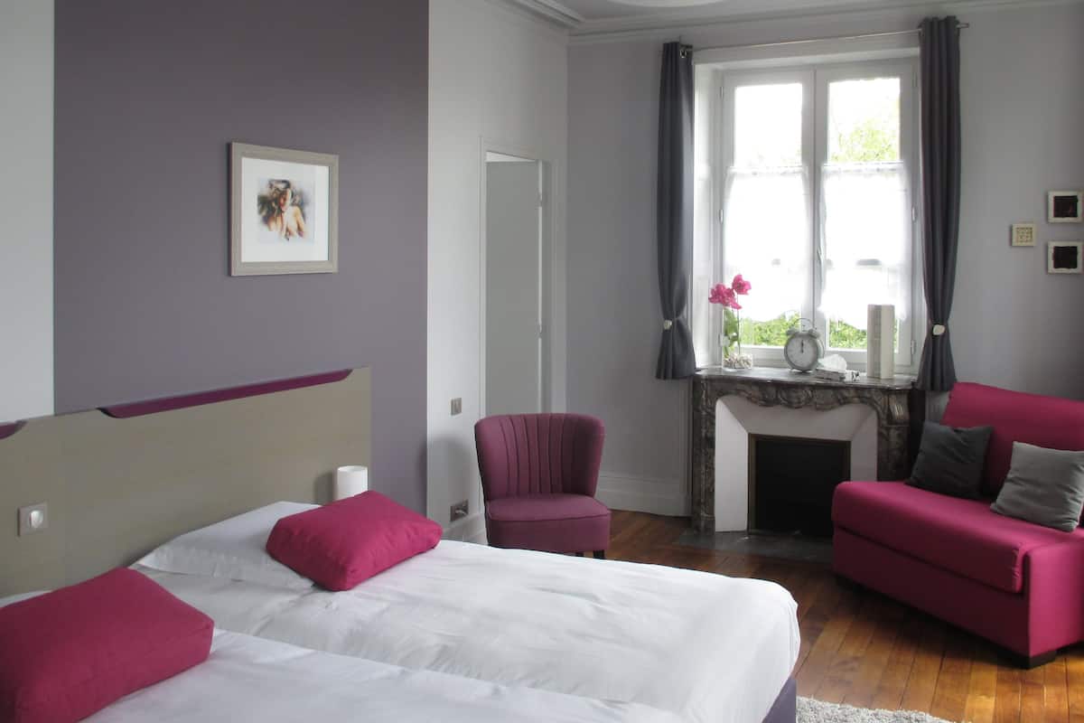 A bedroom with a bed, two chairs, a fireplace, and a window with curtains.