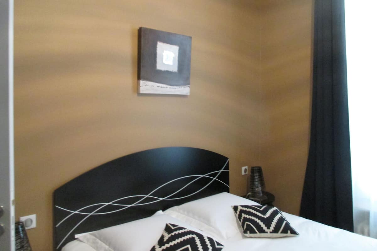 A hotel room with a bed, two pillows, a nightstand, a wall-mounted picture, and a curtain.