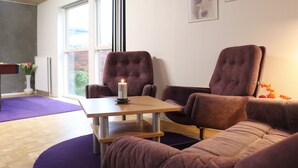 Una sala de estar moderna con una mesa de madera, dos sillones morados y un sofá morado. Una vela encendida está sobre la mesa, y hay obras de arte enmarcadas en la pared.