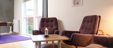 Een moderne woonkamer met een houten tafel, twee paarse fauteuils en een paarse bank. Op de tafel staat een brandende kaars, en aan de muur hangen kunstwerken in lijsten.