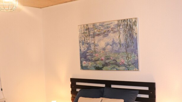Un dormitorio con techo de madera, cama con colcha azul oscuro, dos lámparas de noche y un cuadro de paisaje en la pared.