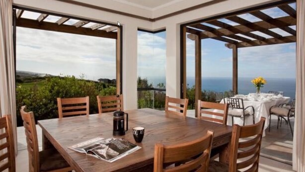 Un espace repas avec une table et des chaises en bois, de grandes fenêtres offrant une vue sur la mer et une structure en forme de pergola.