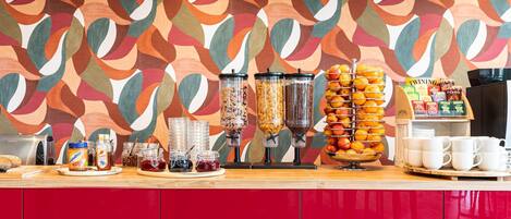 Una barra de desayuno con diversas opciones de comida, una máquina de café y un muro abstracto y colorido.