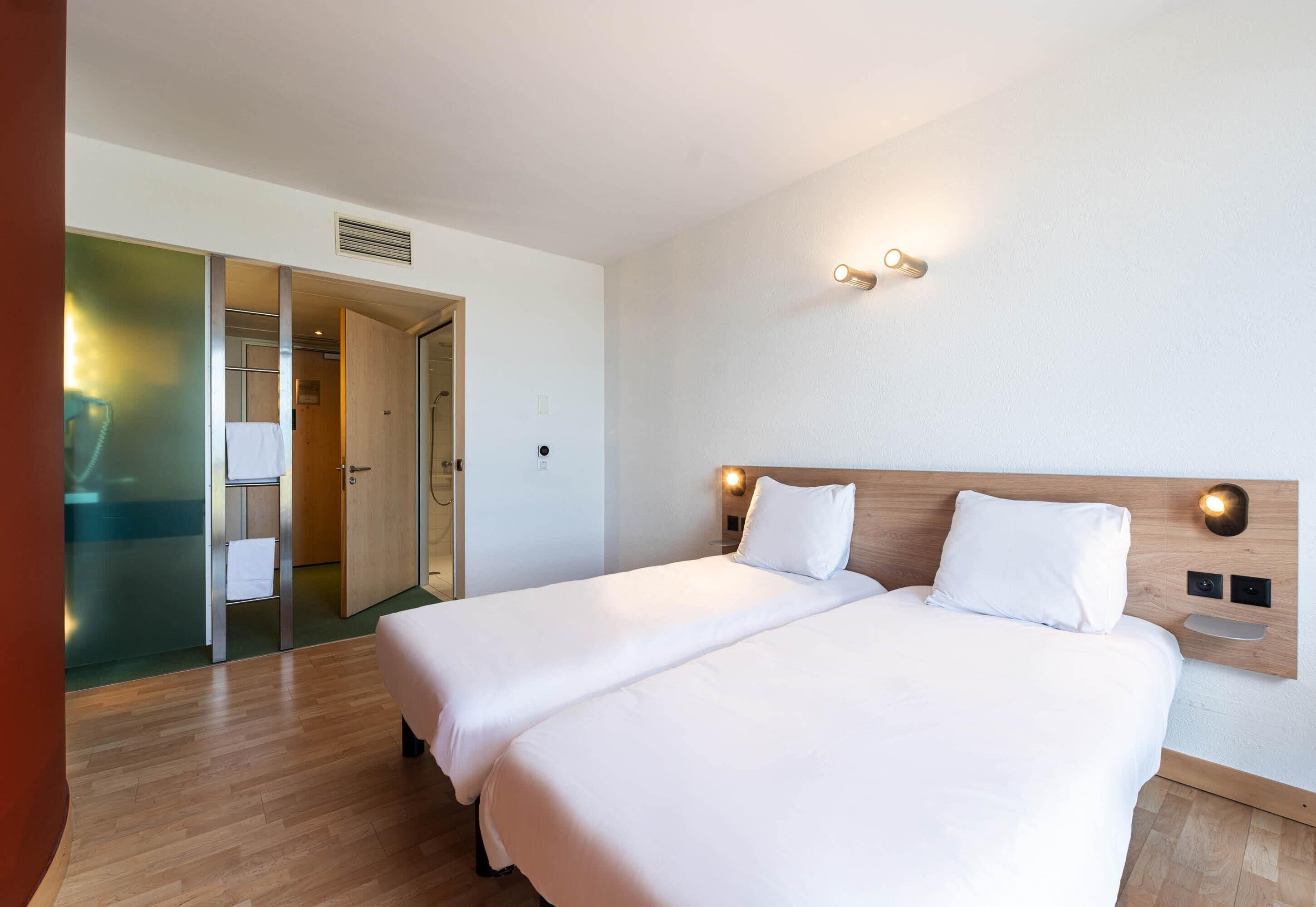 Habitación de hotel con dos camas, cabecero de madera, luces fijadas a la pared y puerta corredera hacia un baño.